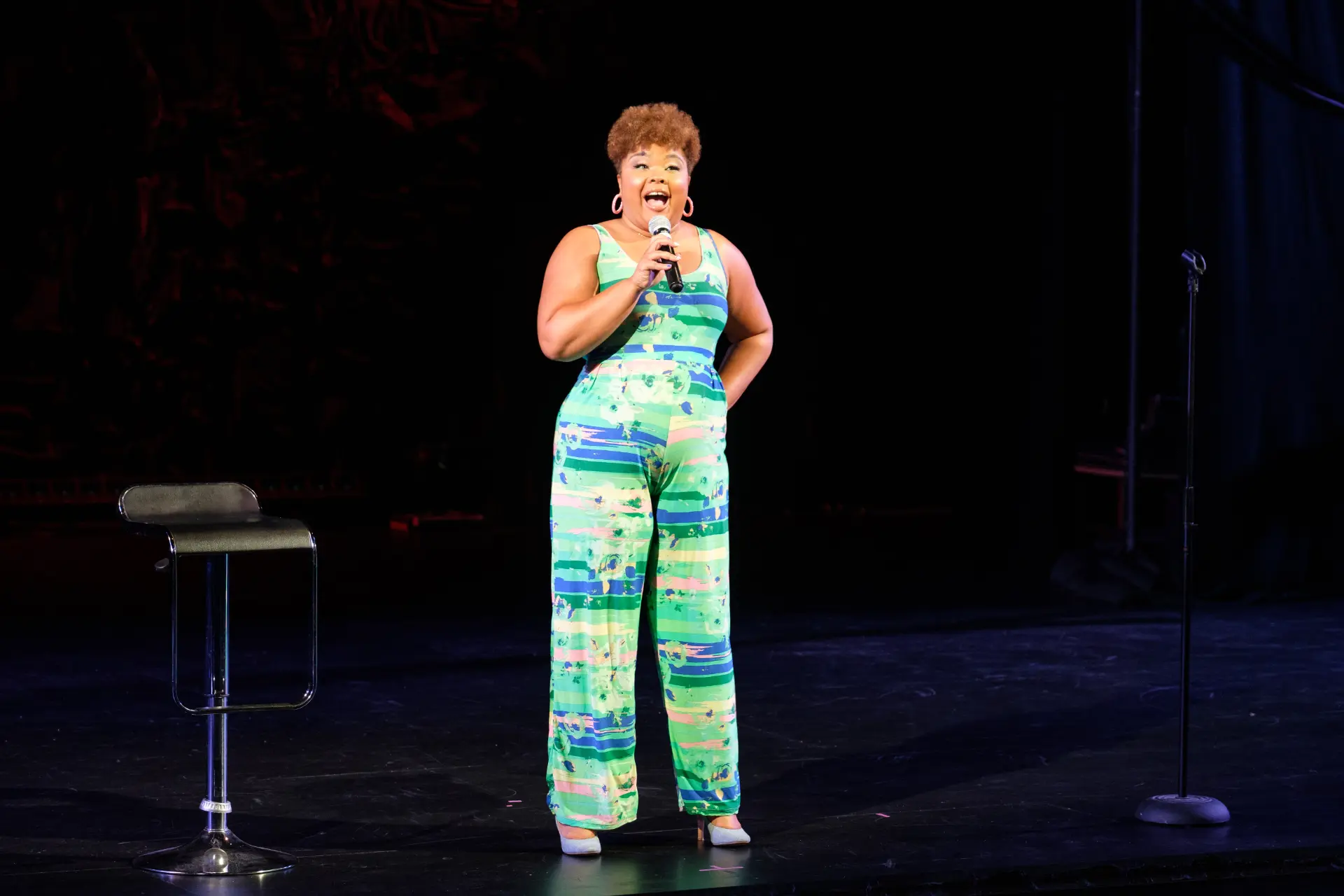 Diana Veiga telling a story onstage in a green patterned jumpsuit
